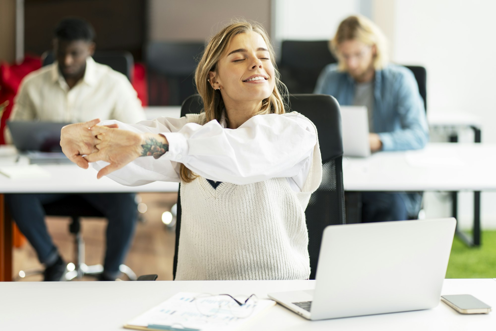 Young, happy businesswoman stretching at her desk in busy office